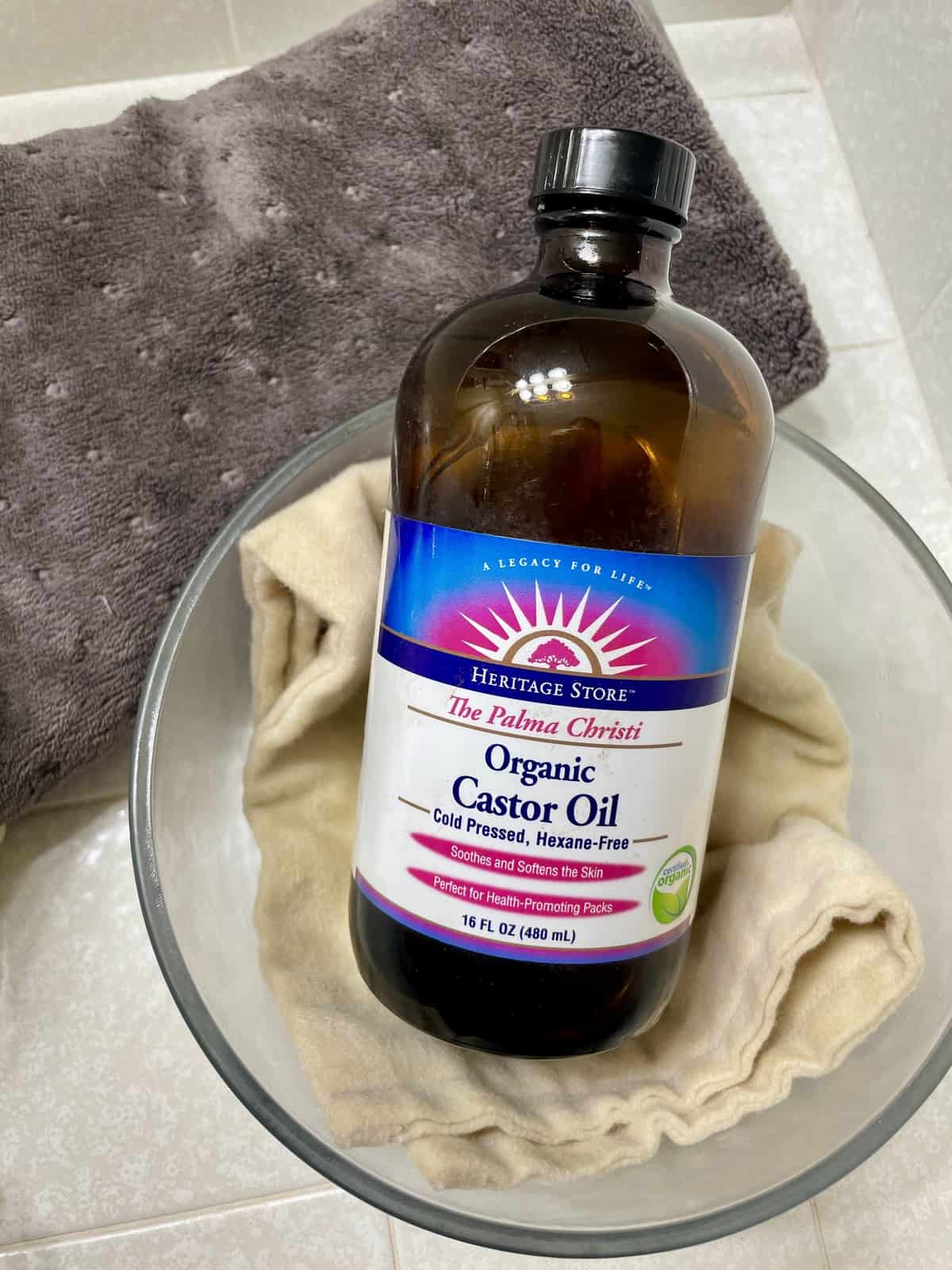 A bottle of organic castor oil sitting inside a glass container with an organic cotton cloth and a heating pad in the background